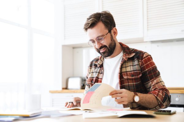 Portrait of excited bearded man wearing eyeglasses smiling while working with notes at home